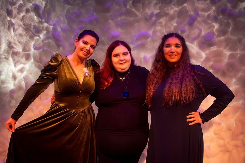 three female figures stand arm in arm in front of a cloud backdrop